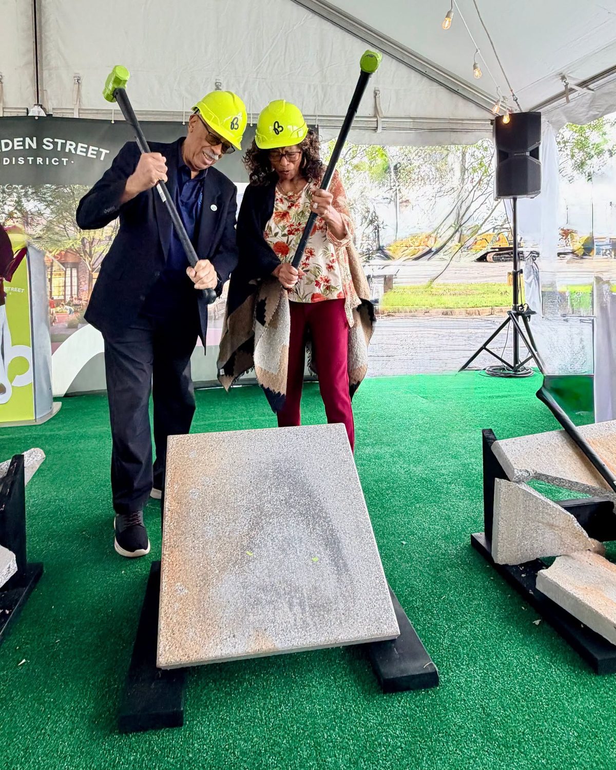 Two guests in hard hats swing sledgehammers at a concrete slab under the Bolden Street District groundbreaking tent