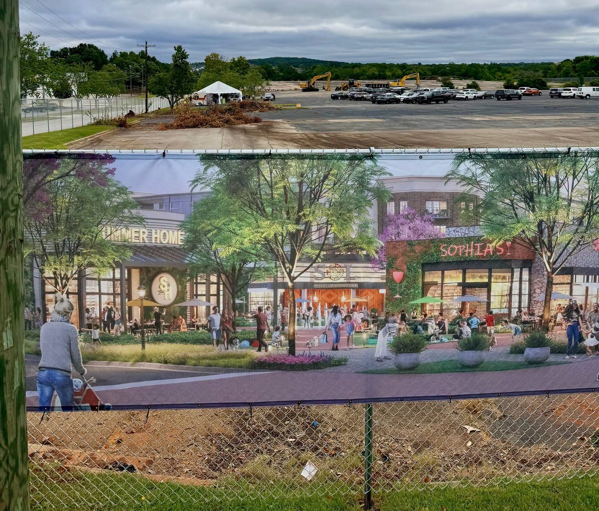A rendering banner of shops and cafes on the construction fence, with the empty former Sam's Club lot and excavators behind it