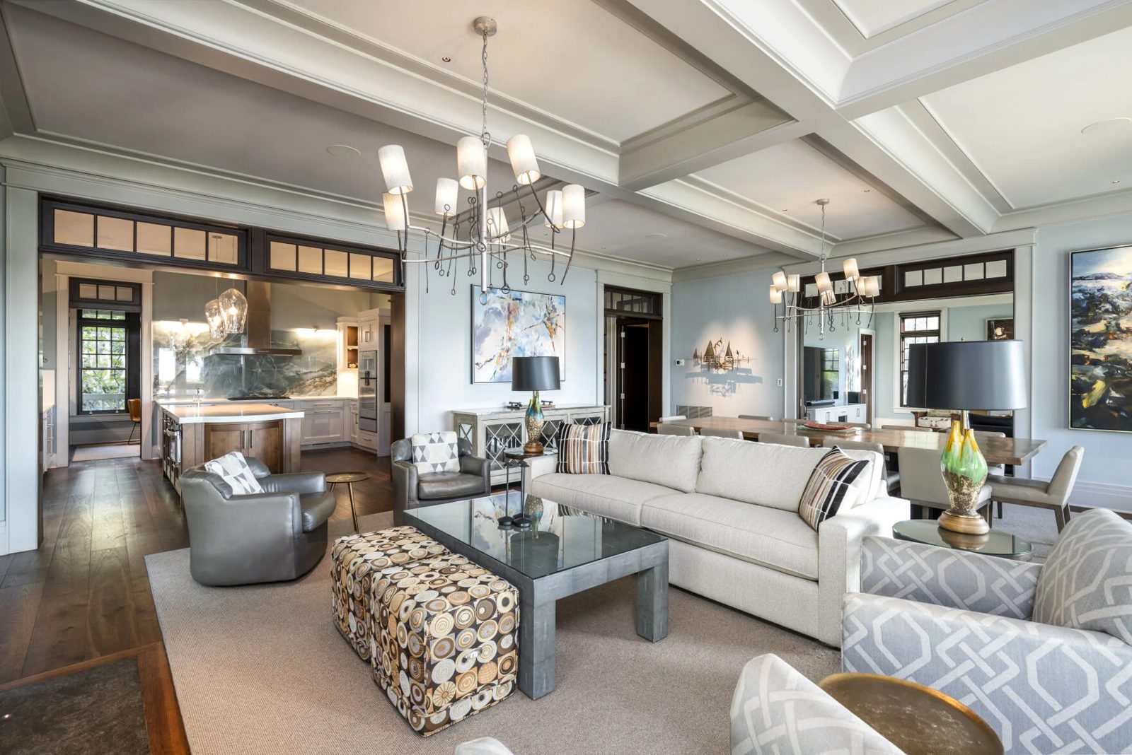A living room under a coffered ceiling, open to a kitchen with a stone backsplash