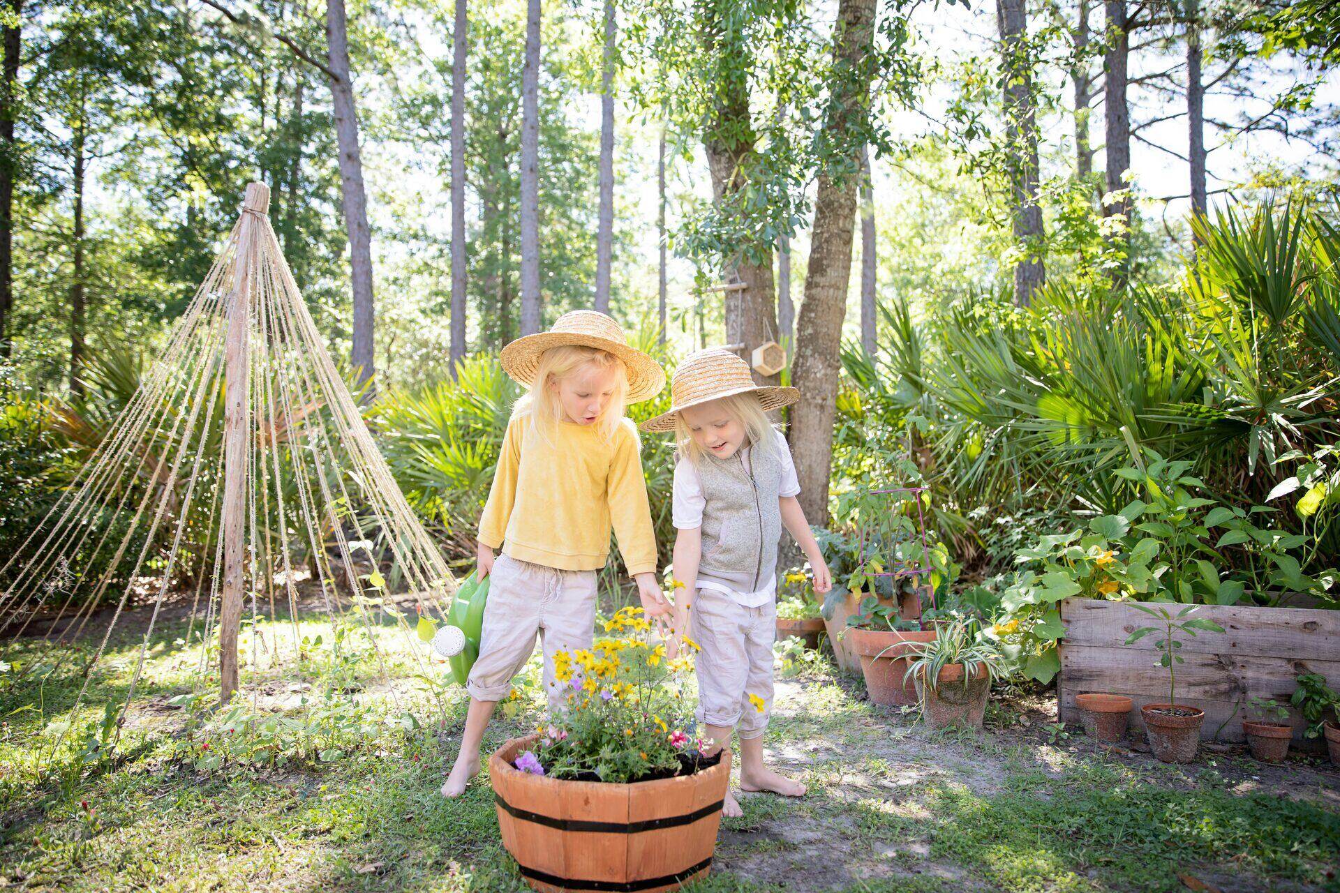 Two children in sun hats among potted flowers and pines on a summer afternoon